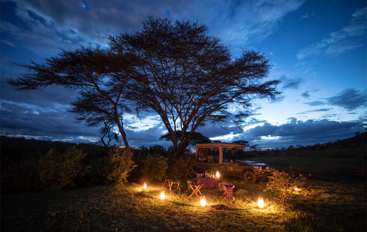 A beautifully lit bush dinner setup under an acacia tree with a safari jeep in the Masai Mara at twilight - Mara Nyika
